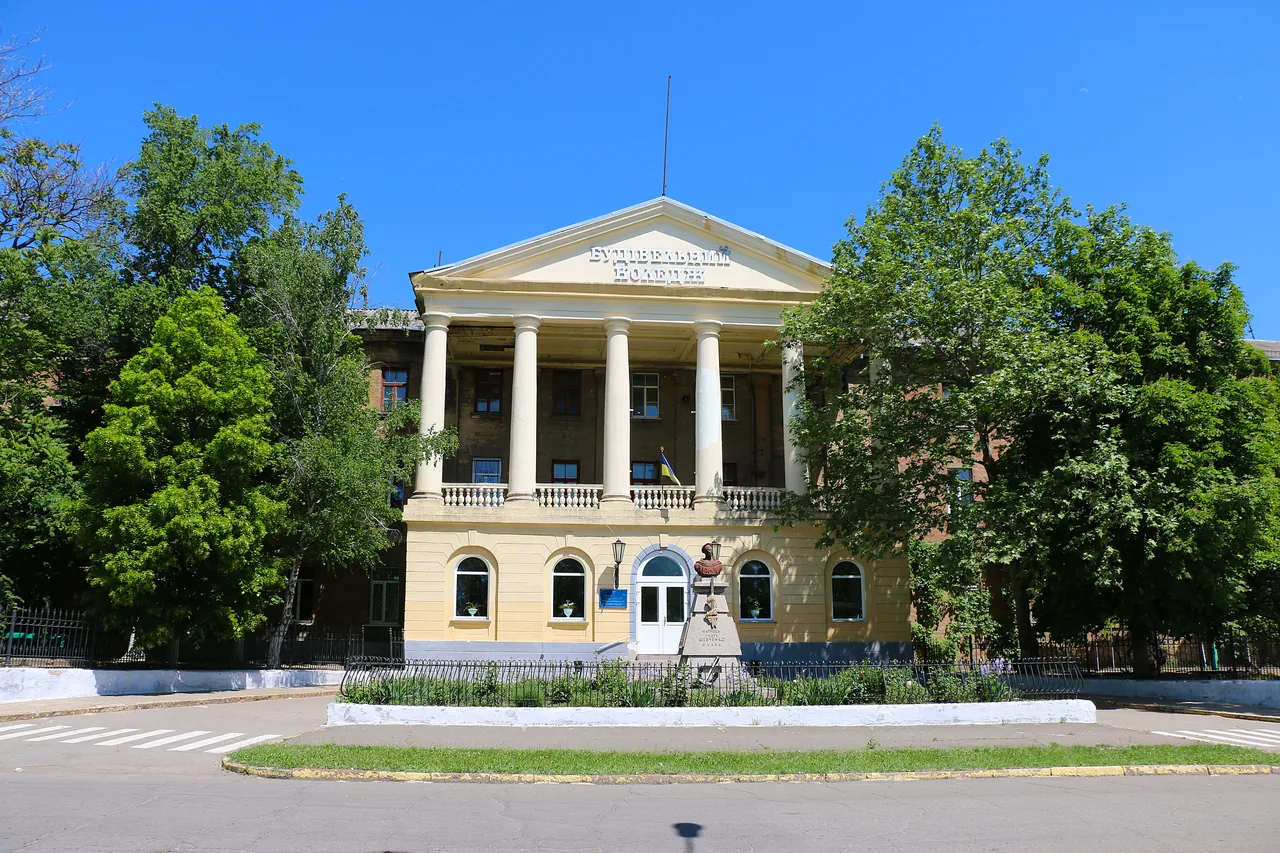 Зелений фронтальний вигляд головного фасаду Миколаївського будівельного коледжу з колонадою, написом на фронтоні та пам’ятником перед входом.