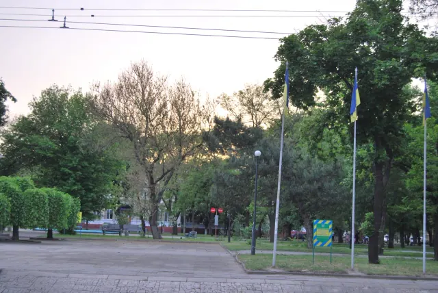 Літній вигляд Аркасівського скверу з алеєю, зеленими деревами, українськими прапорами й охоронним щитом.