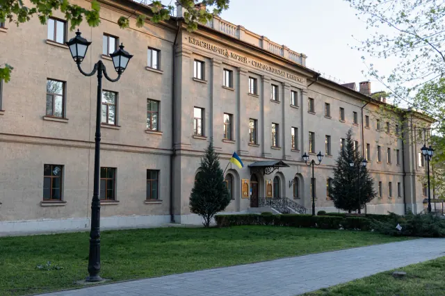 Весняний вигляд повного фасаду Миколаївського обласного краєзнавчого музею у будівлі Старофлотських казарм із написом, центральним входом і зеленим подвір’ям.
